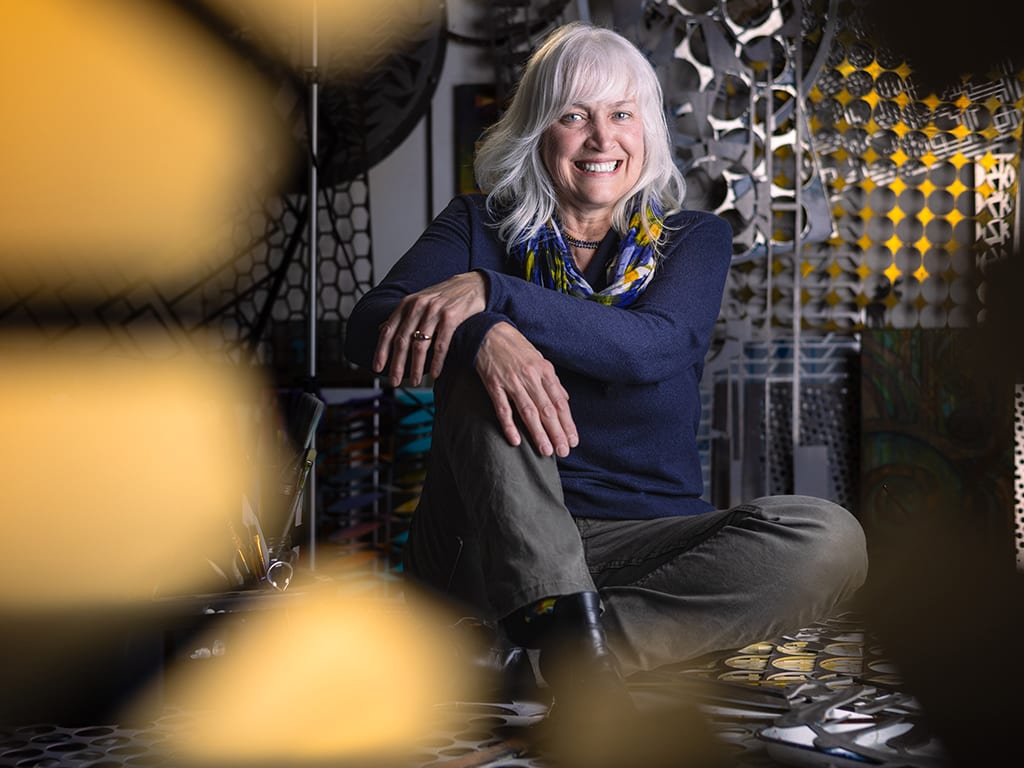 Portrait of Lynn standing at her workbench with lots of laser cut metal shapes around her.
