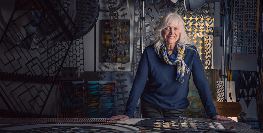 Lynn wearing blue sweater and scarf. Standing with hands resting on her workbench. She is smiling and surrounded by large metal shapes.